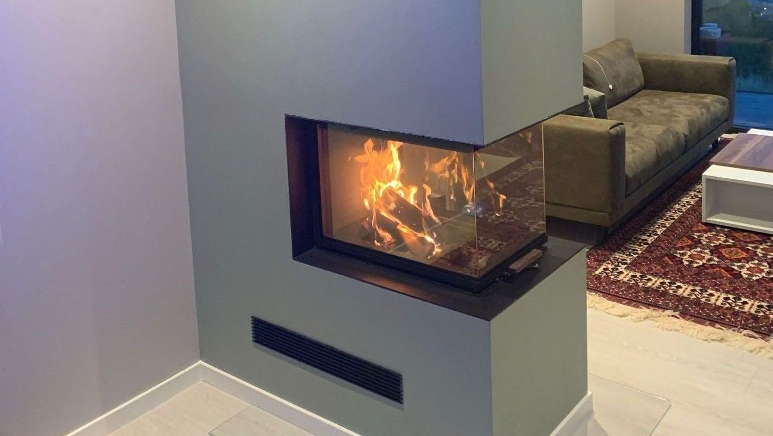 Moderne peis med glass på hjørnet og synlige flammer, plassert i et stilrent og lyst oppholdsrom.